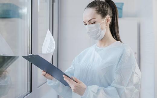A gowned, masked clinician reviews a chart on a clipboard in a clinical setting