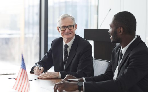 Two officials in business suits at a conference table with a U.S. flag, one signing a document