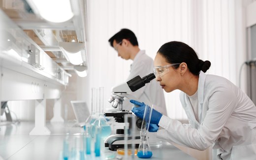 Two researchers in a laboratory, one examining a sample under a microscope while a colleague works at a fume hood in the background