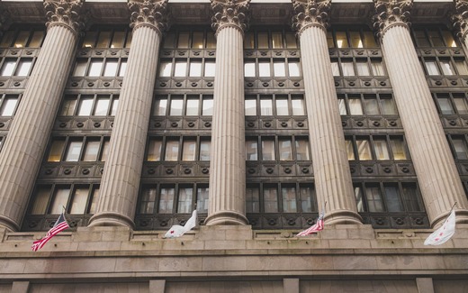 A row of Corinthian columns rises along the stone facade of a government office building flying American flags