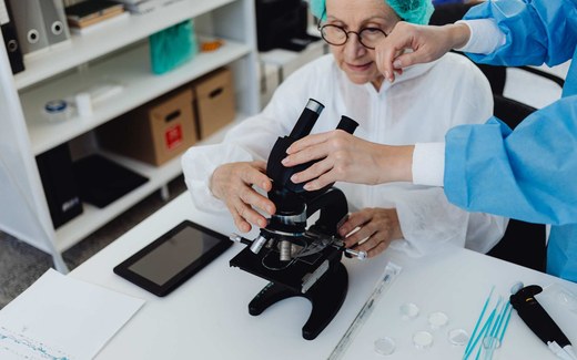 A senior scientist in a cleanroom gown and hairnet adjusts a microscope while a gowned colleague assists at the bench