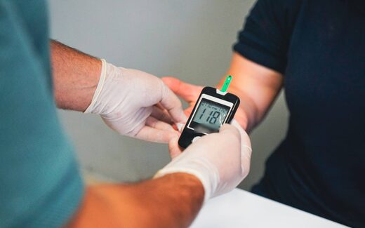 A clinician's gloved hands hold a glucose meter to a patient's fingertip, a generic stand-in for City Therapeutics' anticoagulation research