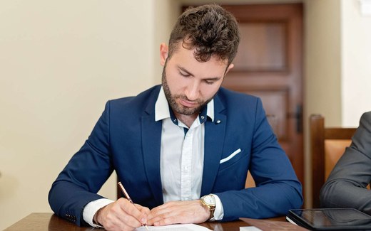 An executive in a navy suit signs an official document at a wooden desk
