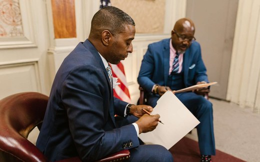 Two men in suits seated with an American flag behind them, one writing on a document held on a clipboard while the other reviews papers