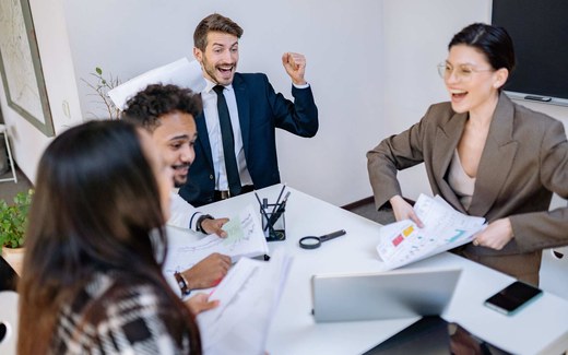 A team of colleagues in business attire celebrate around a conference table with charts and papers