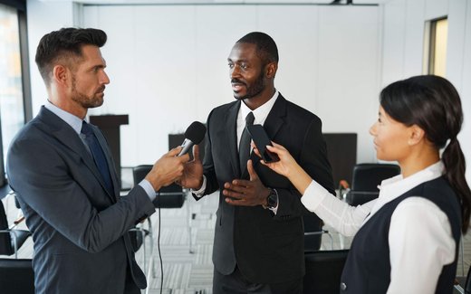 Three people in business attire speak into microphones held out by reporters in a conference room