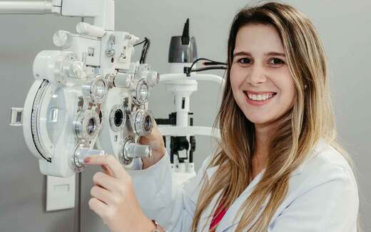 A smiling optometrist points to a phoropter during a vision exam in a bright eye clinic