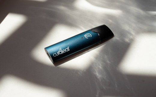 Curaleaf International's QMID inhaler, a slim blue-green metallic device with a dark mouthpiece and the Curaleaf leaf mark, lying on white fabric in dappled sunlight