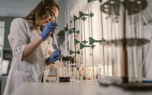 A researcher in a white lab coat and safety glasses uses a pipette over laboratory glassware