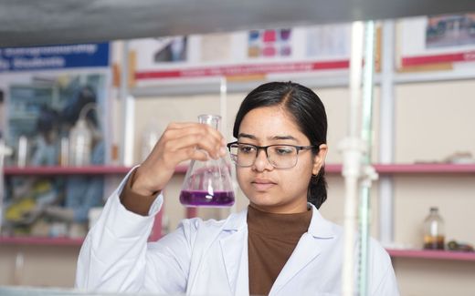A researcher in a lab coat and glasses examines a flask of purple liquid held up to the light