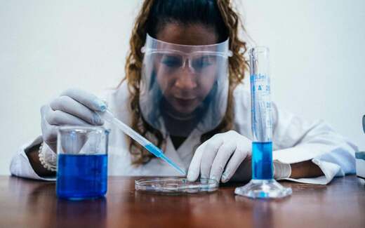A researcher in a lab coat and face shield uses a pipette with blue reagent, standing in for small-molecule drug research