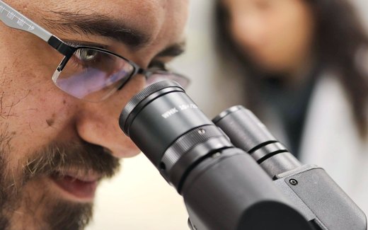A researcher wearing glasses looks through a binocular microscope while a colleague in a lab coat stands in the background
