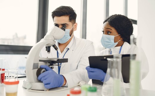 Two masked lab researchers review notes on a clipboard while operating a microscope, surrounded by lab glassware