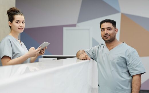 Two clinicians in scrubs confer at a bright hospital reception desk