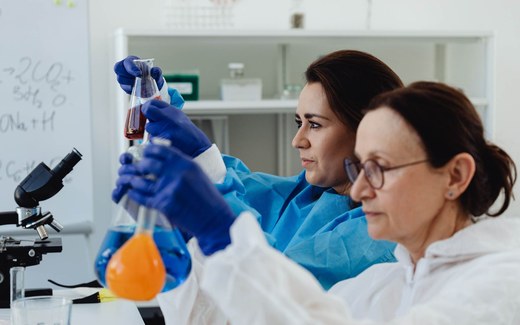 Two researchers in white and blue lab coats examine chemical flasks on a laboratory bench