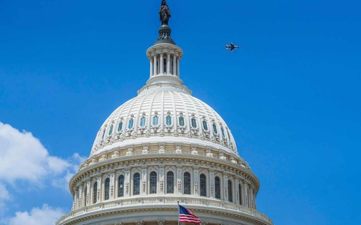 The dome of the United States Capitol against a clear blue sky, with a jet aircraft passing high to its right