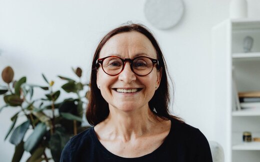A smiling woman wearing glasses in a bright home living room, a generic stand-in for a patient managing care at home