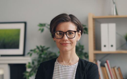 A professional wearing glasses and a dark blazer in an office with bookshelves and plants in the background