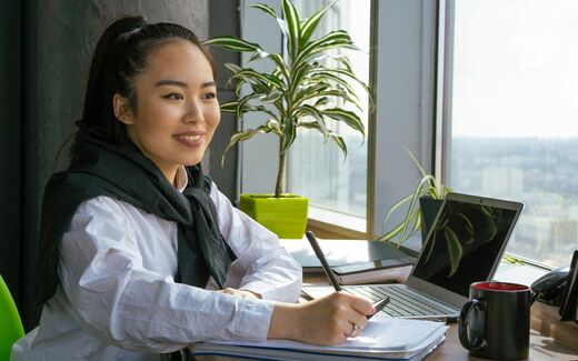 A professional reviews documents and a laptop at a bright office desk by a window