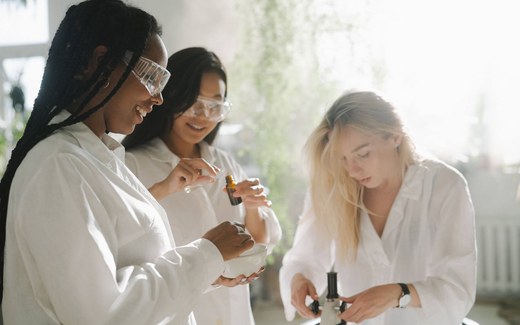 Three scientists in a sunlit lab examine a bottle and a bowl of solution while adjusting laboratory equipment