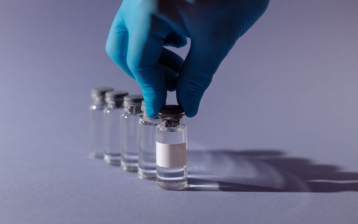 A gloved hand lifts a glass vial from a row of vials on a laboratory bench, its label left blank