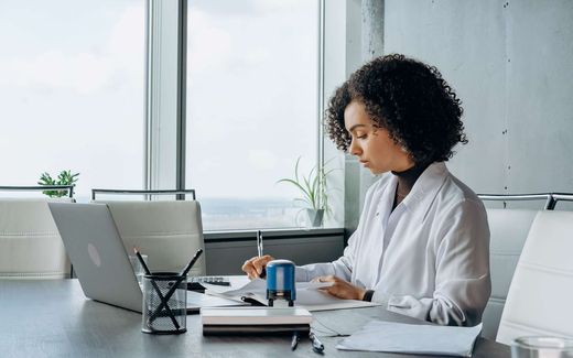 A professional in a white blouse signs and stamps documents at a desk beside a laptop