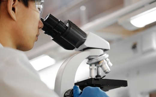 A researcher wearing safety glasses and a white lab coat looks through a microscope eyepiece while adjusting a sample slide