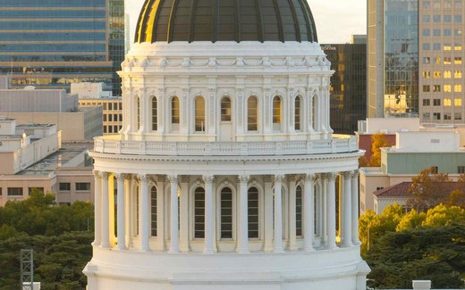 A government building dome with classical columns rises above a city skyline at sunset