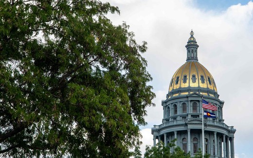 A gold-domed capitol building with an American flag rises above green trees against a partly cloudy sky