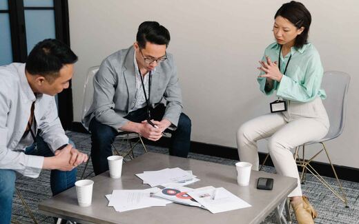 Three colleagues with lanyards review printed documents around a table