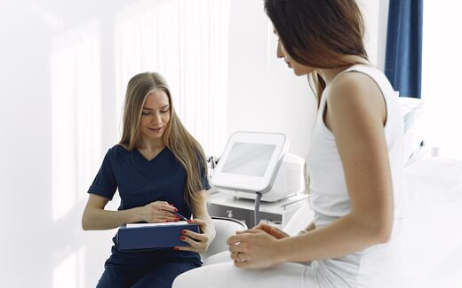 A clinician in scrubs consults with a patient beside a bedside monitor