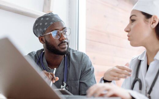 Two clinicians in scrubs and a lab coat discussing a case over a laptop by a bright window, a generic stand-in for clinical and regulatory coordination