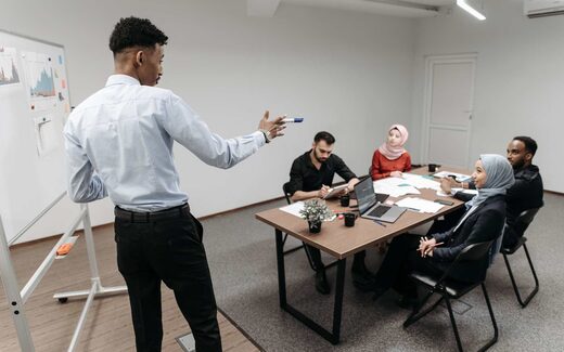 A colleague gestures toward a whiteboard while briefing a seated team