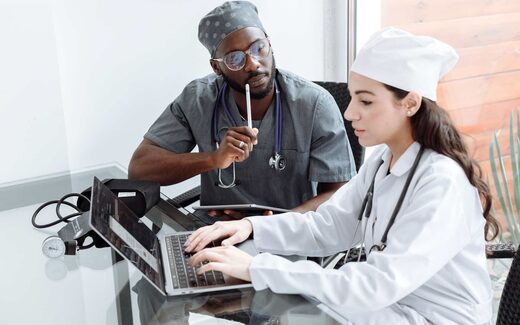 Two clinicians in scrubs review patient data together on a laptop