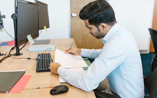 A man at a desk with two monitors annotates a printed document with a pen
