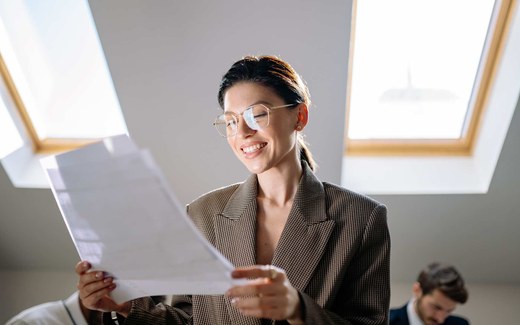A smiling professional in glasses and a tweed blazer reads a printed document by a sunlit skylight window