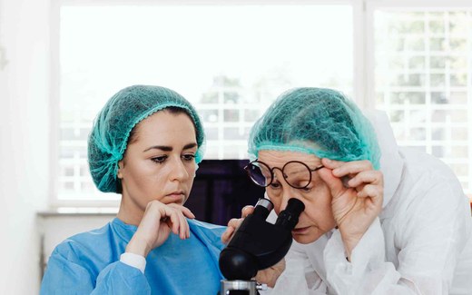 Two gowned, hairnet-capped laboratory researchers examining a sample under a microscope, one adjusting her glasses to look into the eyepiece