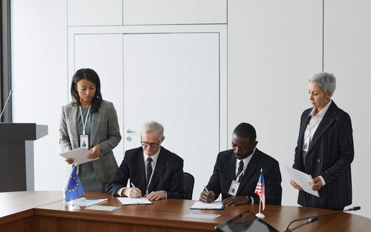 Two officials sign documents at a table bearing European Union and American flags while colleagues review papers standing behind them