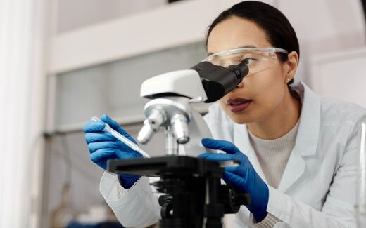 A scientist in a lab coat and safety glasses examines a sample through a microscope while holding a pipette