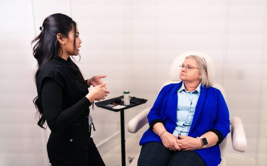 A clinician in a dark top speaks with a seated patient during a consultation in a bright exam room