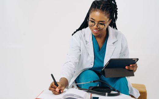 A physician in a white coat and scrubs writes in a notebook beside a stethoscope and a tablet