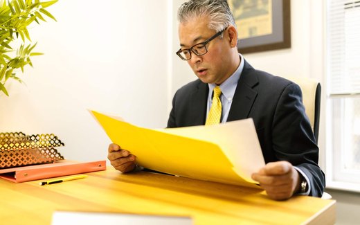 A man in a dark suit reads intently from an open yellow folder at a wooden desk