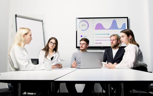 A team reviews performance charts on a wall-mounted display during a meeting around a conference table