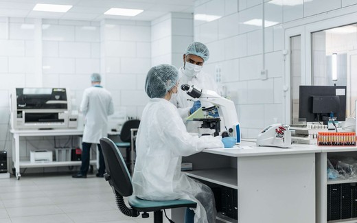 Two lab technicians in cleanroom garb examining a sample under a microscope in a bright diagnostics laboratory