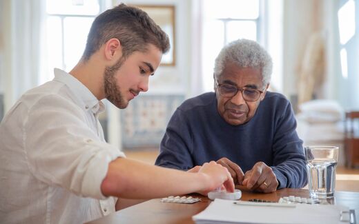 A younger man helps an older man sort prescription medication into a daily pill organizer at a table