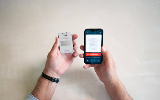 A hand holds the credit-card-sized HeartBeam ECG device beside a smartphone running the HeartBeam app