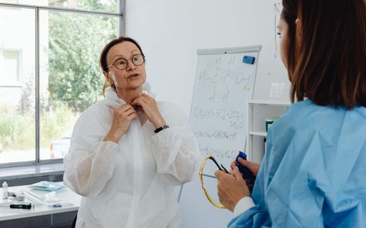 A senior scientist in a protective coverall speaks with a colleague in a lab beside a whiteboard covered in chemical notation