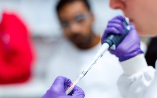 A gloved researcher holds a pipette over a sample tube while a colleague looks on in a laboratory