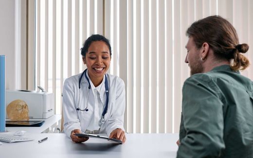 A physician in a white coat and stethoscope reviews a tablet with a patient during a clinic consultation