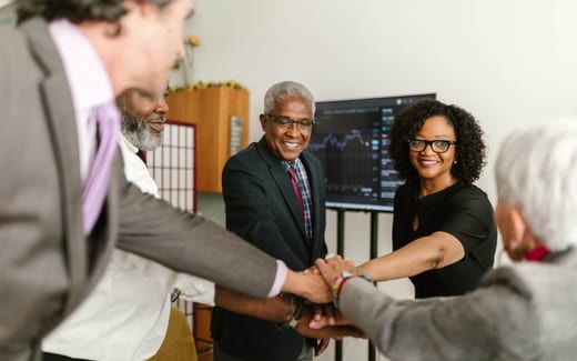 A team stacks hands together in celebration in an office, with a market chart visible on a screen behind them